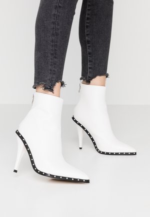 Personne portant des bottines blanches à talons hauts pointus avec bordure noire cloutée et jean skinny noir effiloché.