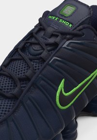 Κοντινό πλάνο αθλητικού παπουτσιού Nike Shox σε ναυτικό μπλε, με μαύρες και νέον πράσινες πινελιές, που δείχνει τα κορδόνια, το πλεκτό πάνω μέρος και το κεντημένο λογότυπο Nike.