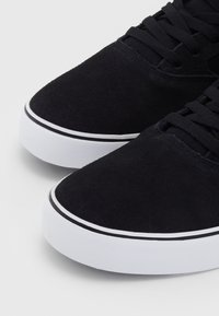 Czarne zamszowe sneakersy z zaokrąglonym noskiem, teksturowaną cholewką i białą gumową podeszwą z czarnym paskiem wzdłuż krawędzi. Z wiązanym sznurowaniem.