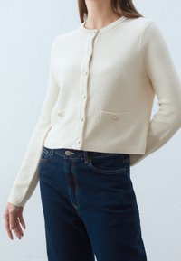 Krämfärgad virkade cardigan med ett texturerat mönster, rund hals, knappstängning och två framfickor; i kombination med mörkblå jeans.