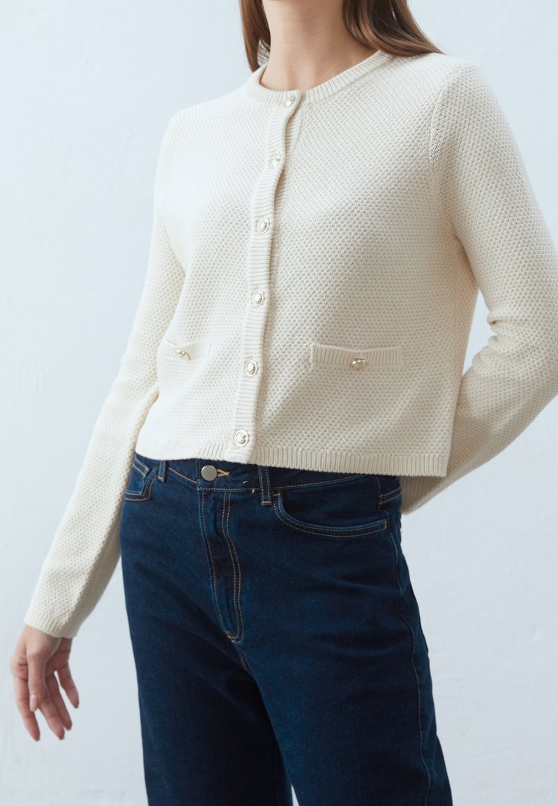 Krämfärgad virkade cardigan med ett texturerat mönster, rund hals, knappstängning och två framfickor; i kombination med mörkblå jeans.