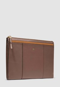 Bruine leren documentenhouder met ritssluiting, voorzien van een beige accentstrook en een klein metalen logo aan de voorkant, rechtopstaand tegen een grijze achtergrond.