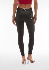 Femme portant un pantalon noir ajusté et des sandales à talons hauts transparentes, vue de dos sur un fond blanc.