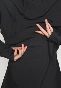 Mani che tirano su il retro di una giacca nera per rivelare una tasca nascosta orizzontale.