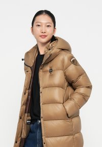 Hnedá puffa bunda s kapucňou, s prešívaným dizajnom, so zipsovým uzáverom a logom na rukáve. Nosí sa cez čierny top.
