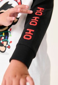 Langarmshirt mit einem schwarzen Ärmel und rotem "HO HO HO"-Text, weißem Körper und buntem Charakterdesign, aus Baumwollstoff.