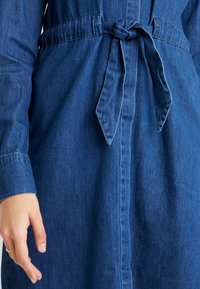 Abito di jeans con vita regolabile tramite cordoncino. Presenta maniche lunghe, una chiusura frontale con bottoni e un ricco colore blu con tessuto testurizzato.