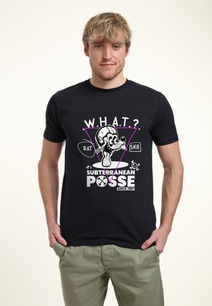 Młody mężczyzna ubrany w czarny t-shirt z kreskówkową myszą w hełmie, napis „W.H.A.T.?“, „Subterranean Posse“ i „Since 1967“, ręce w kieszeniach.
