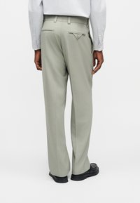 Άνδρας που φοράει ελαφριά γκρι παντελόνια ραμμένα στα μέτρα και μαύρα παπούτσια, στέκεται με την πλάτη προς την κάμερα μπροστά από μια απλή φόντο.