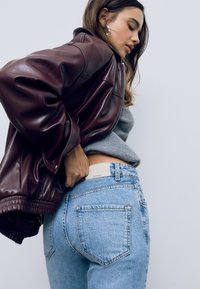 Bordová kožená bomber bunda, šedý sveter, svetlomodré vysoké džínsy s viditeľnou značkou, prezentujúce ležérny vrstvený outfit.