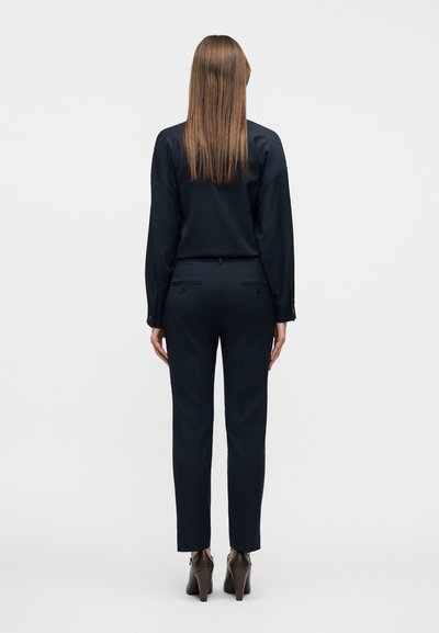 Γυναίκα που φαίνεται από πίσω φορώντας μαύρο μακρυμάνικο τοπ, μαύρο παντελόνι προσαρμοσμένης γραμμής και καφέ μποτάκια με τακούνι σε λευκό φόντο.