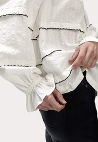 Blusa bianca a trama con bordo scallop nero, con polsini arricciati e design a strati, indossata su pantaloni aderenti scuri.