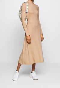 Beige pitkähihainen ribbimekko, jossa on korkea pääntie, virtaava siluetti, ja se on yhdistetty nahan kaltaiseen käärmeentäplikkääseen laukkuun sekä valkoisiin lenkkikenkiin.