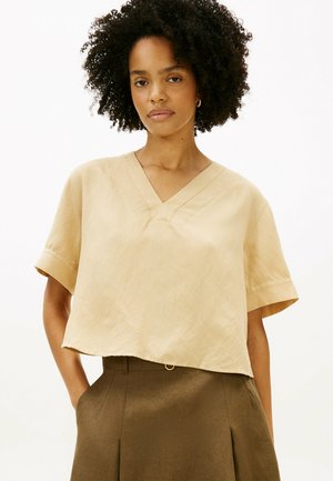 Frau mit lockigem Haar, die eine beige V-Ausschnitt-Bluse mit kurzen Ärmeln und hoch taillierte braune Hose trägt, steht mit den Händen in den Taschen.