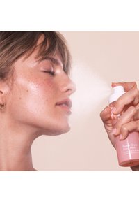 Bouteille de spray rose étiquetée "Brume Éclat Pêche" tenue près du visage, brume se dispersant vers la peau. Design lisse et épuré avec un embout blanc.