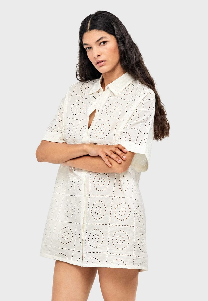 Robe chemise blanche à manches courtes avec col à boutons, présentant des motifs circulaires découpés et une coupe ample. Texture de tissu léger.