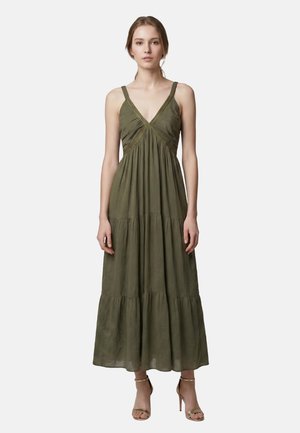 Jeune femme debout portant une robe longue sans manches de couleur olive avec des fines bretelles et des sandales dorées à talons hauts sur un fond uni.