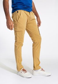 Bežové cargo kalhoty s bočními kapsami, slim fit a mírně zúženým nohavicím. Kombinované s bílými teniskami a modrou košilí.