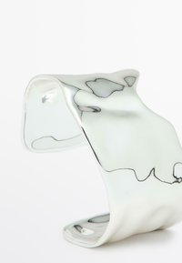 Metalarmband i cuff-stil med en vågig, polerad silveryta och svarta linjedetaljer, som har mjuka kurvor och en öppen design.