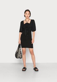 Vestido preto com mangas bufantes e laço na frente; alças finas. Combinado com uma bolsa preta em rede e sandálias planas pretas. Fundo claro e simples.