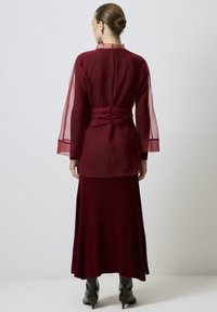 Conjunto en capas color granate con mangas largas y transparentes y detalle fruncido en la espalda. La falda es acanalada y fluye hasta el tobillo, combinada con botas negras.