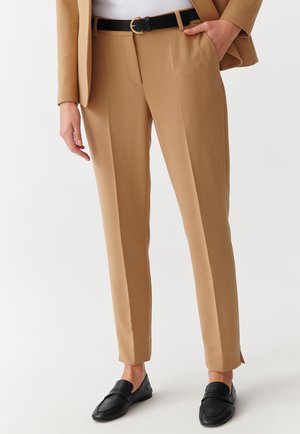 Beige tailored trousers made from a smooth fabric, featuring a slim fit, front pleats, and side slits, paired with black loafers.