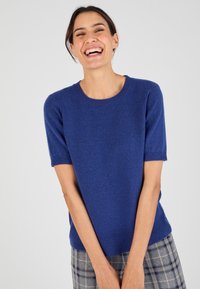 Pull-over en tricot bleu à manches courtes avec un col rond ; texture douce et bords côtelés ; porté par-dessus un pantalon à carreaux gris.