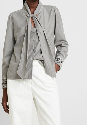 Personne portant un chemisier rayé noir et blanc avec un col noué et des poignets brodés de fleurs, associé à un pantalon blanc.