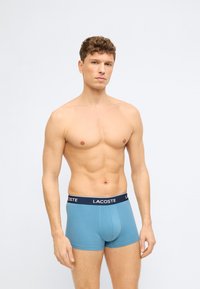 Άνδρας με κοντά κατσαρά μαλλιά που φοράει μπλε εσώρουχα Lacoste boxer, στέκεται μπροστά σε απλό φωτεινό φόντο, κοιτάζοντας μπροστά με ουδέτερη έκφραση.