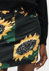 Μαύρη τζιν φούστα με μεγάλα κίτρινα prints ηλιοτροπίων, φορεμένη με μαύρο πλεκτό πουλόβερ, το χέρι ακουμπάει κοντά στην πλαϊνή τσέπη της φούστας.