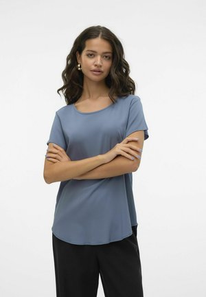 Short-sleeve top in soft blue fabric with a rounded neckline and a curved hem. Worn with dark pants, showcasing a relaxed fit.