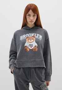 Hoodie grigio corto con grafica di un orso che indossa una maglietta con la scritta "New York". Abbinato a pantaloni della tuta grigi con la scritta "B".