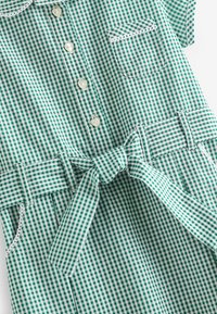 Vihreä ja valkoinen ruudullinen mekko, jossa on lyhyet hihat, napit edessä, tasku ja solmittava vyötärö. Valmistettu kevyestä kankaasta, jossa on sileä tekstuuri.