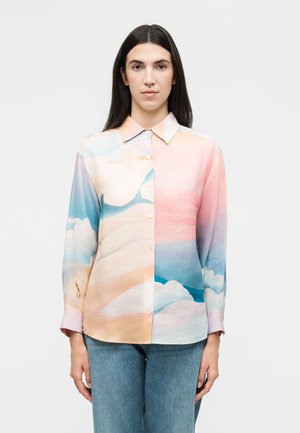 Γυναίκα με μακριά σκούρα μαλλιά φοράει πουκάμισο παστέλ με μοτίβα από απαλά σύννεφα και ουρανό, σε συνδυασμό με ανοιχτό μπλε τζιν.