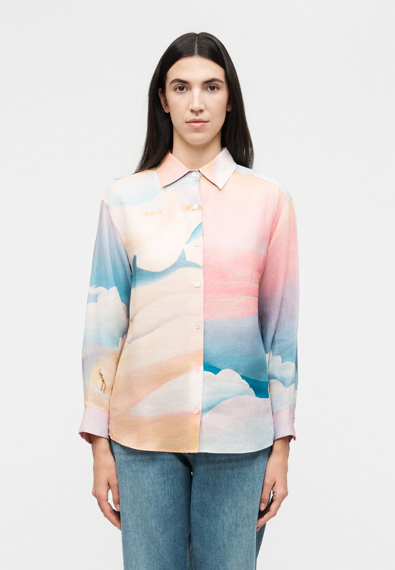 Γυναίκα με μακριά σκούρα μαλλιά φοράει πουκάμισο παστέλ με μοτίβα από απαλά σύννεφα και ουρανό, σε συνδυασμό με ανοιχτό μπλε τζιν.