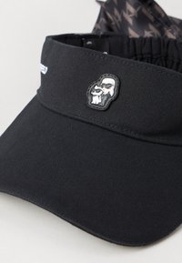 Czarna czapka z daszkiem z teksturowanej tkaniny, z gumowaną łatą przedstawiającą postać i kota. Zawiera elastyczny pasek dla regulowanego dopasowania.
