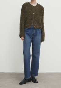 Bruine pluizige cardigan met lange mouwen en ronde hals, voorzien van grote knopen; gecombineerd met blauwe jeans met rechte pijpen en zwarte schoenen.