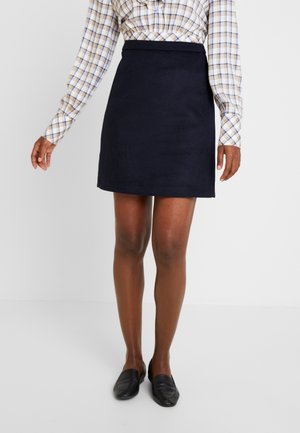 Woman wearing a navy blue mini skirt, plaid long-sleeve shirt, and black loafers, standing against a plain white background.