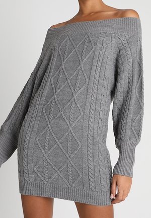 Vrouw draagt een grijze off-shoulder kabelgebreide sweaterjurk met lange mouwen en geribbelde manchetten, zoom en halslijn.