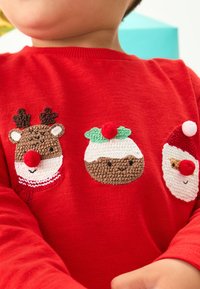 Chemise rouge à manches longues avec trois motifs de Noël brodés : un renne, un pudding de Noël et le Père Noël, chacun avec des accents colorés.