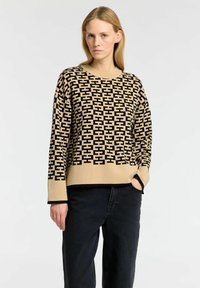 Beige neule, jossa on musta geometrinen kuvio, pyöreä pääntie ja pitkät hihat. Yhdistetty tummiin farkkuihin, joissa on väljä ja rento istuvuus.