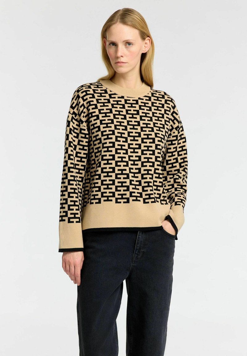 Beige neule, jossa on musta geometrinen kuvio, pyöreä pääntie ja pitkät hihat. Yhdistetty tummiin farkkuihin, joissa on väljä ja rento istuvuus.