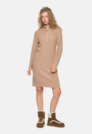 Beiger, strukturierter Strickpulloverkleid mit halbhohem Reißverschlusskragen, langen Ärmeln und schlanker Passform; kombiniert mit braunen, mit Fell gefütterten Knöchelstiefeln.