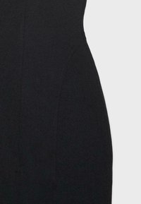 Gros plan d’un vêtement noir avec des coutures verticales visibles formant une taille cintrée sur un fond gris clair uni.