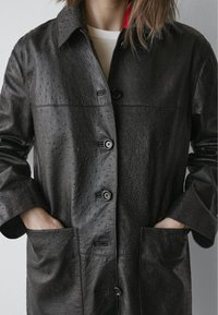 Zwarte leren jas met een getextureerd oppervlak, grote knopen en zakken aan de zijkant. Voorzien van een kraag en een contrasterende voering aan de binnenkant.