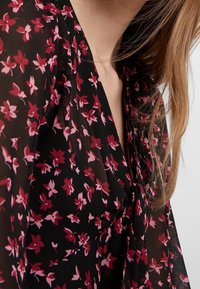 Černá květinová blůza vyrobená z průhledné, lehké látky s růžovými a červenými květinovými vzory. Design obsahuje výstřih do V a volný střih.