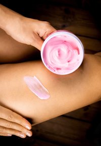 Rosa återfuktande kräm i en rund vit behållare, med en slät klick på bruna hudtoner, som visar en krämig konsistens mot en träbakgrund.