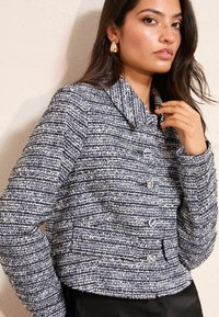 Krótkia kurtka wykonana z niebieskiej i białej tkaniny tweedowej z teksturowanym wzorem, z szerokim kołnierzem i srebrnymi guzikami z przodu.