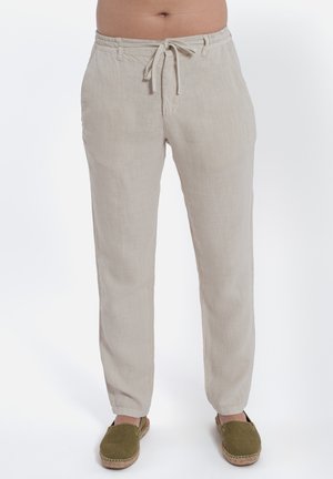 Pantaloni beige in lino con vita con coulisse e gambe affusolate, indossati con espadrillas verdi slip-on su sfondo bianco.