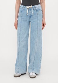 Lys blå vide jeans med snøring i livet og sidelommer, laget av denimstoff med en falmet finish og dobbelt søm detaljer.